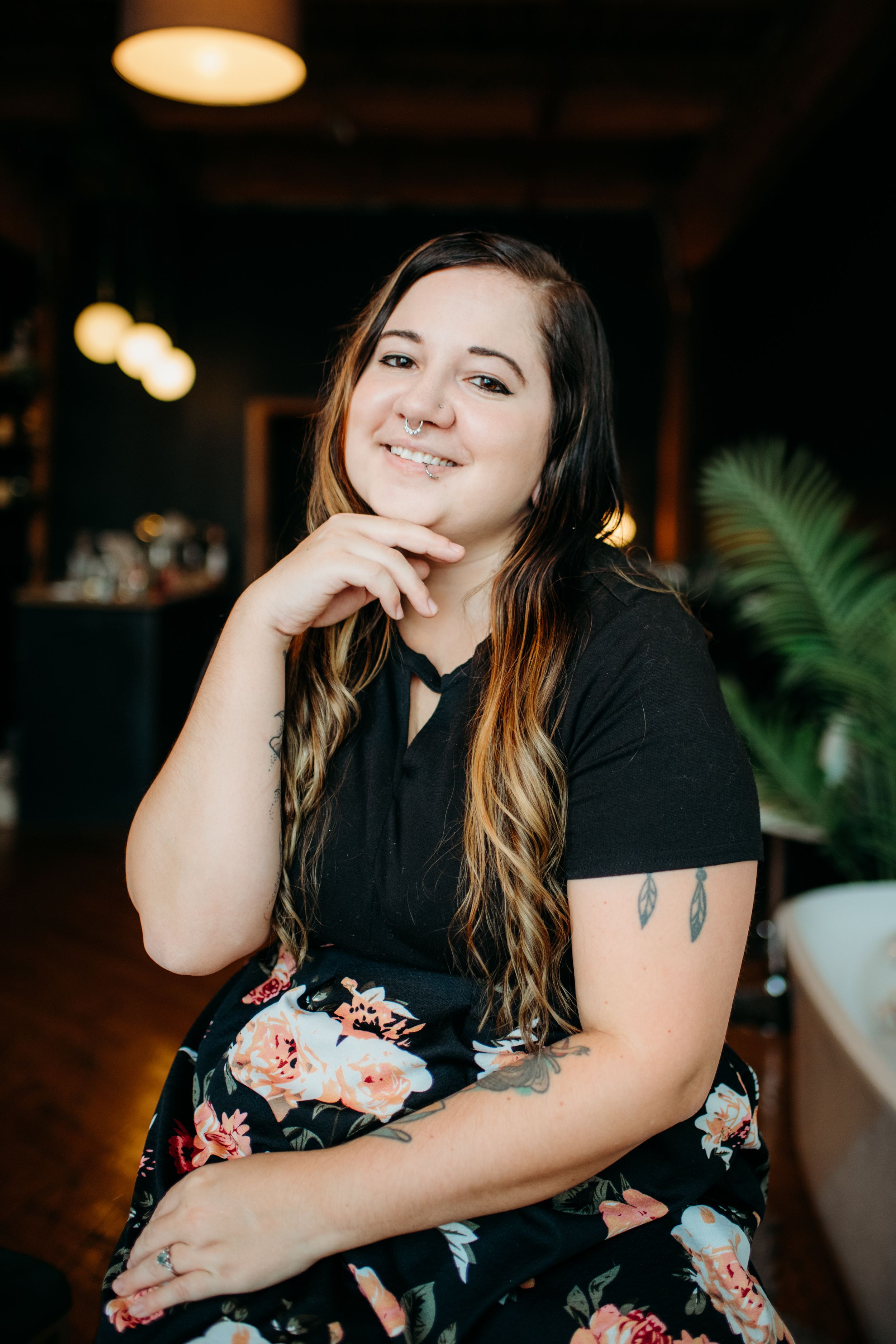 Smiling woman with long hair and tattoos poses in black top and floral skirt in a warmly lit restaurant.