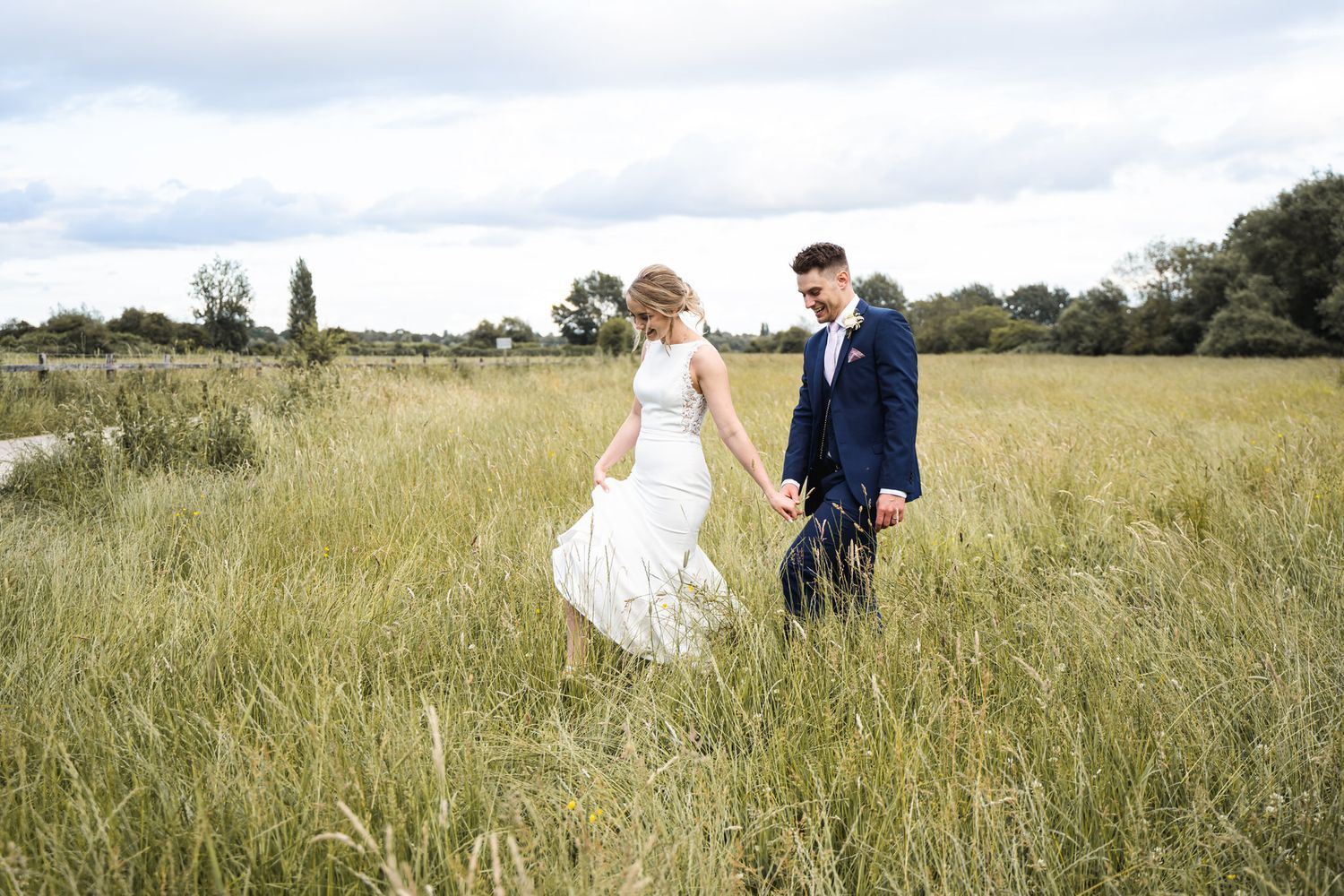Bride and groom walking hand in hand through a lush green field on their wedding day at The Perch Binsey Wedding Venue