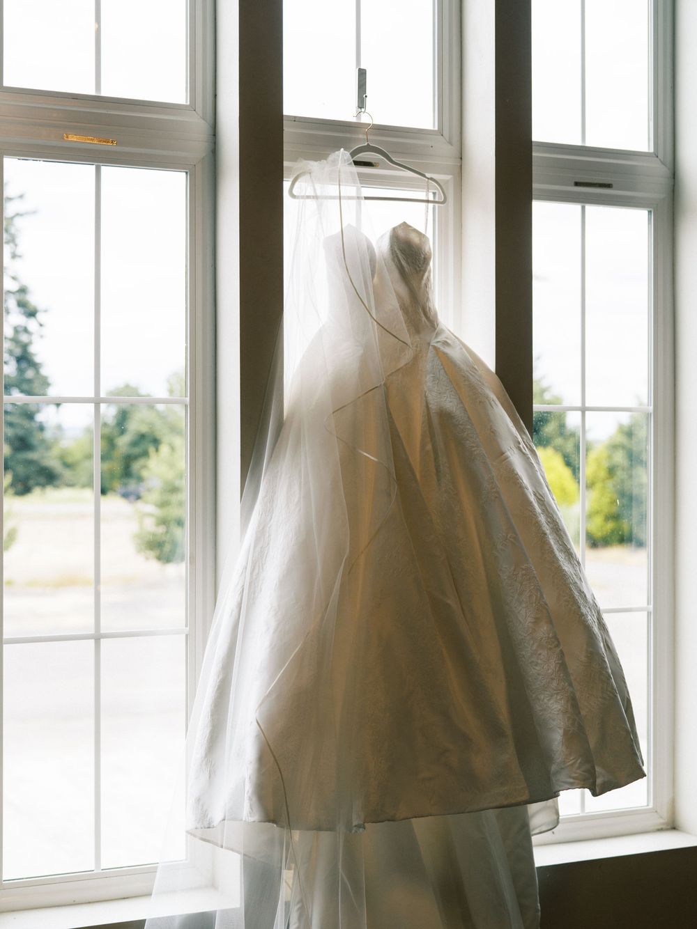 A elegant white wedding dress with veil hanging on a window hanger, backlit by natural light through large windows.