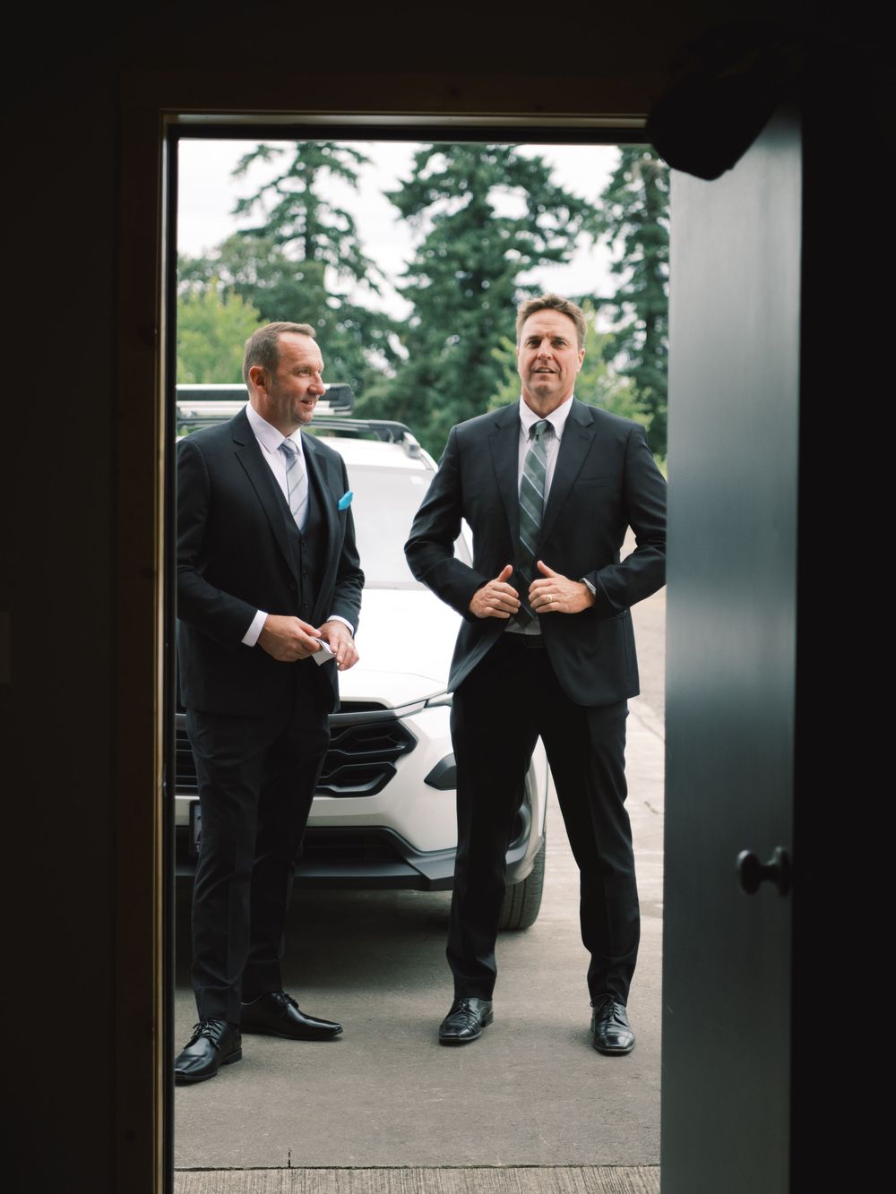Two groomsmen in dark suits smile and laugh outside near a white car on a wedding day.
