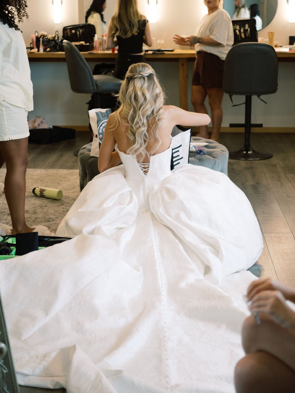 Bride in a voluminous white ball gown with lace-up back sits in a bridal suite during wedding preparations.
