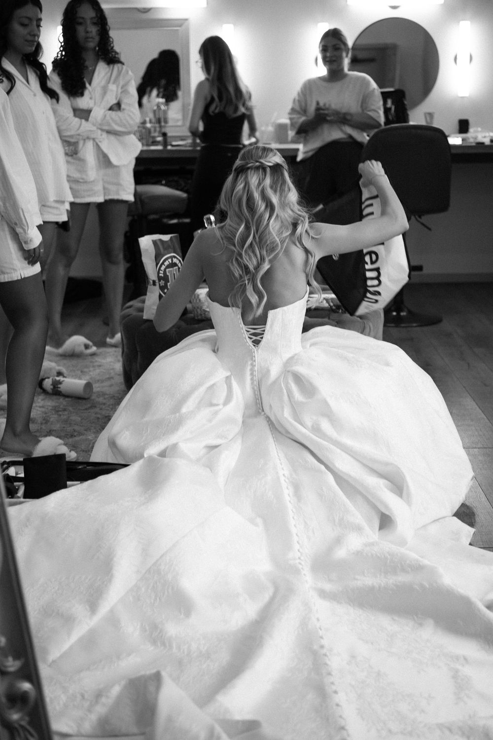 Black and white photo of bride in voluminous ball gown wedding dress being helped by bridesmaids in white robes.