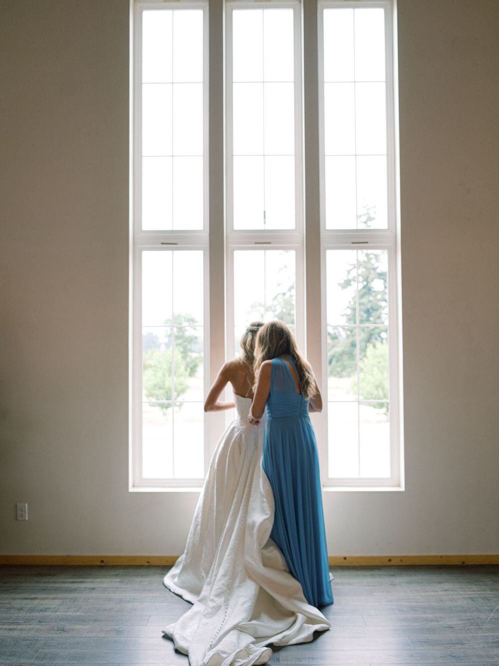 Bride in white gown and bridesmaid in blue dress look out tall windows on wedding day.