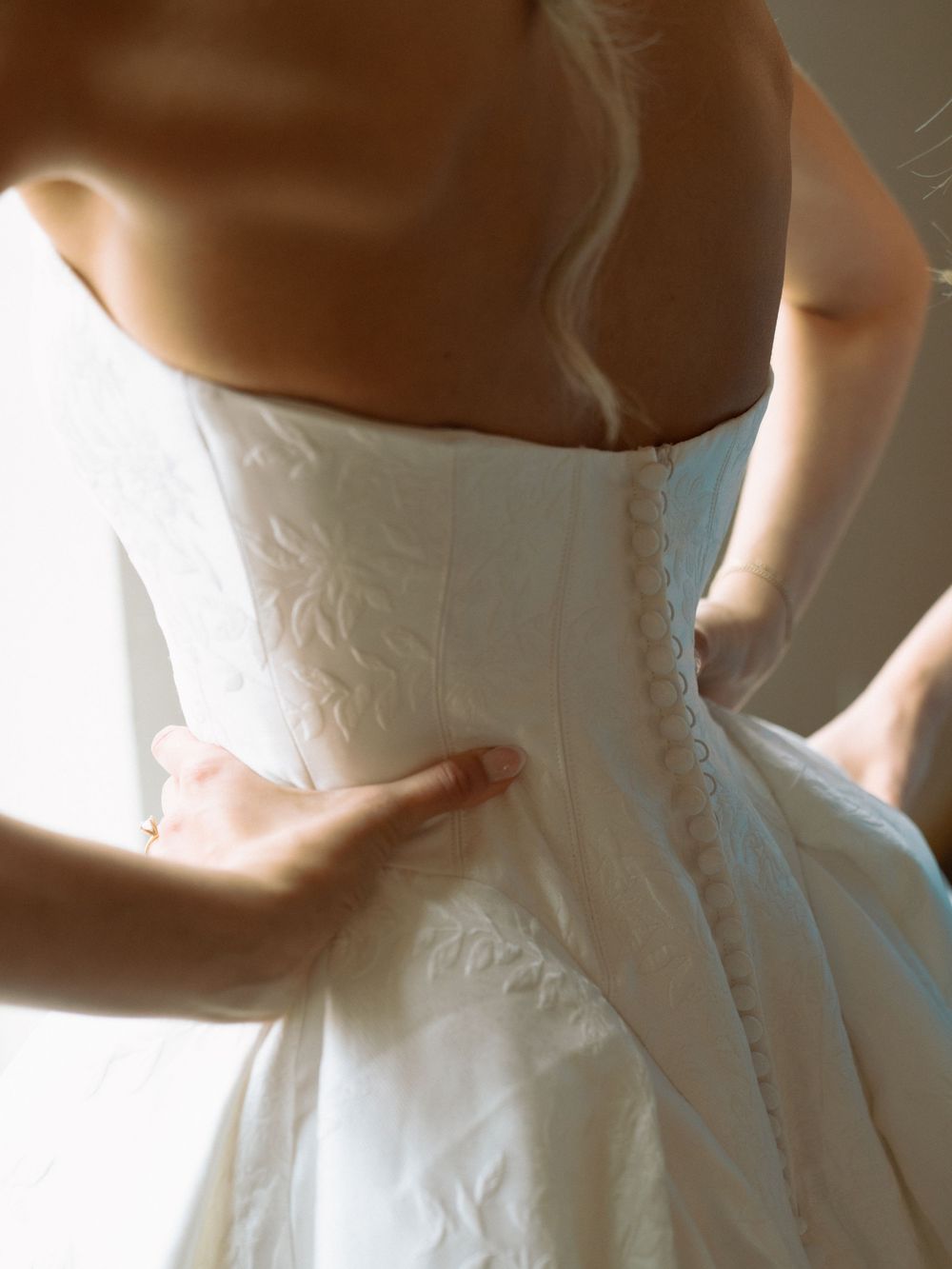 Bride being laced into a white strapless corset wedding gown with floral embroidery.