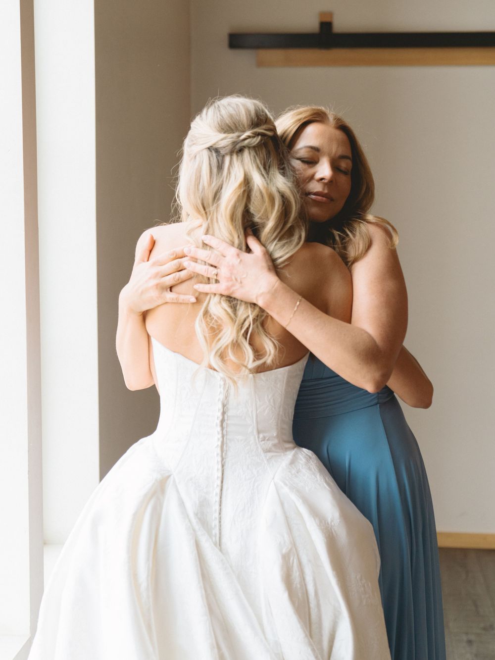 A bride in a white strapless gown embraces a woman in a blue dress during an emotional wedding day moment.