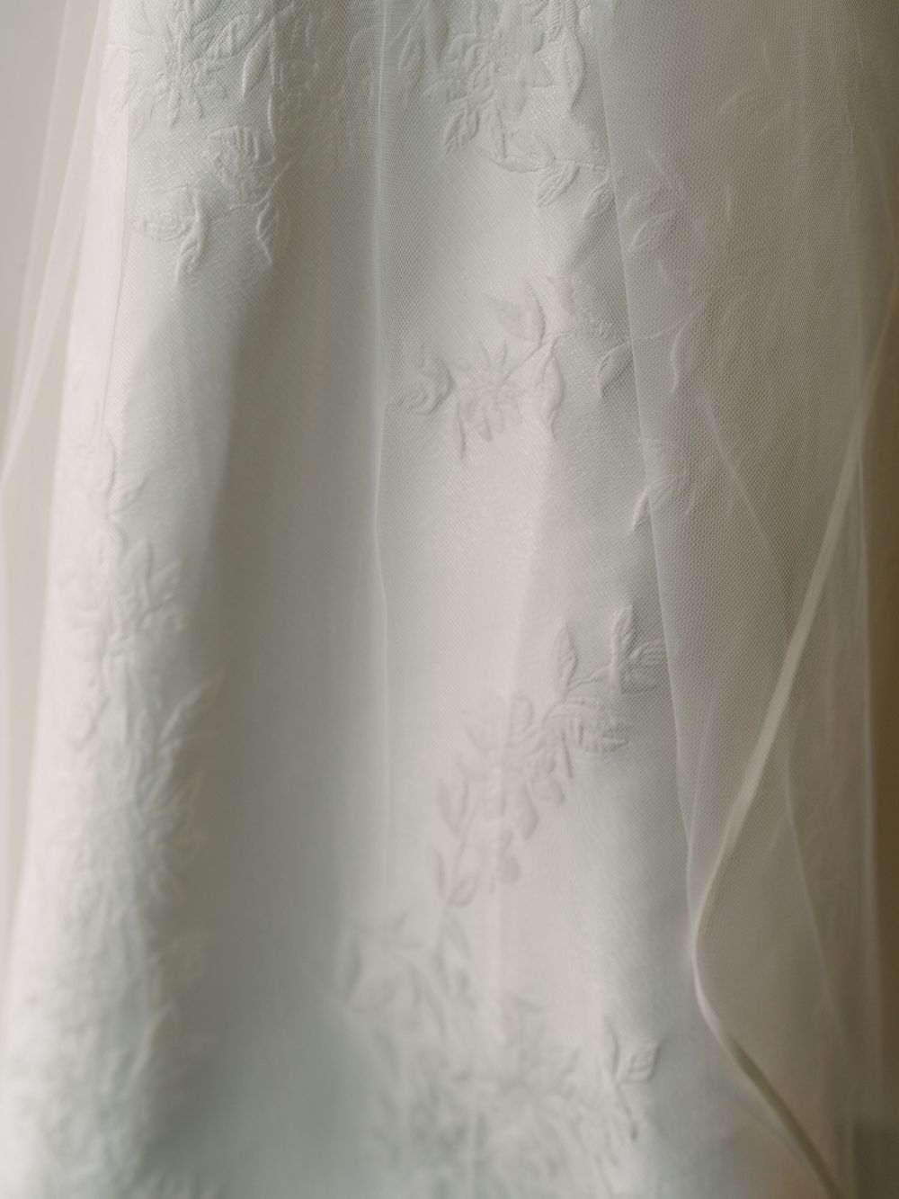 Close-up of white wedding dress fabric with delicate embroidered floral details and sheer overlay.