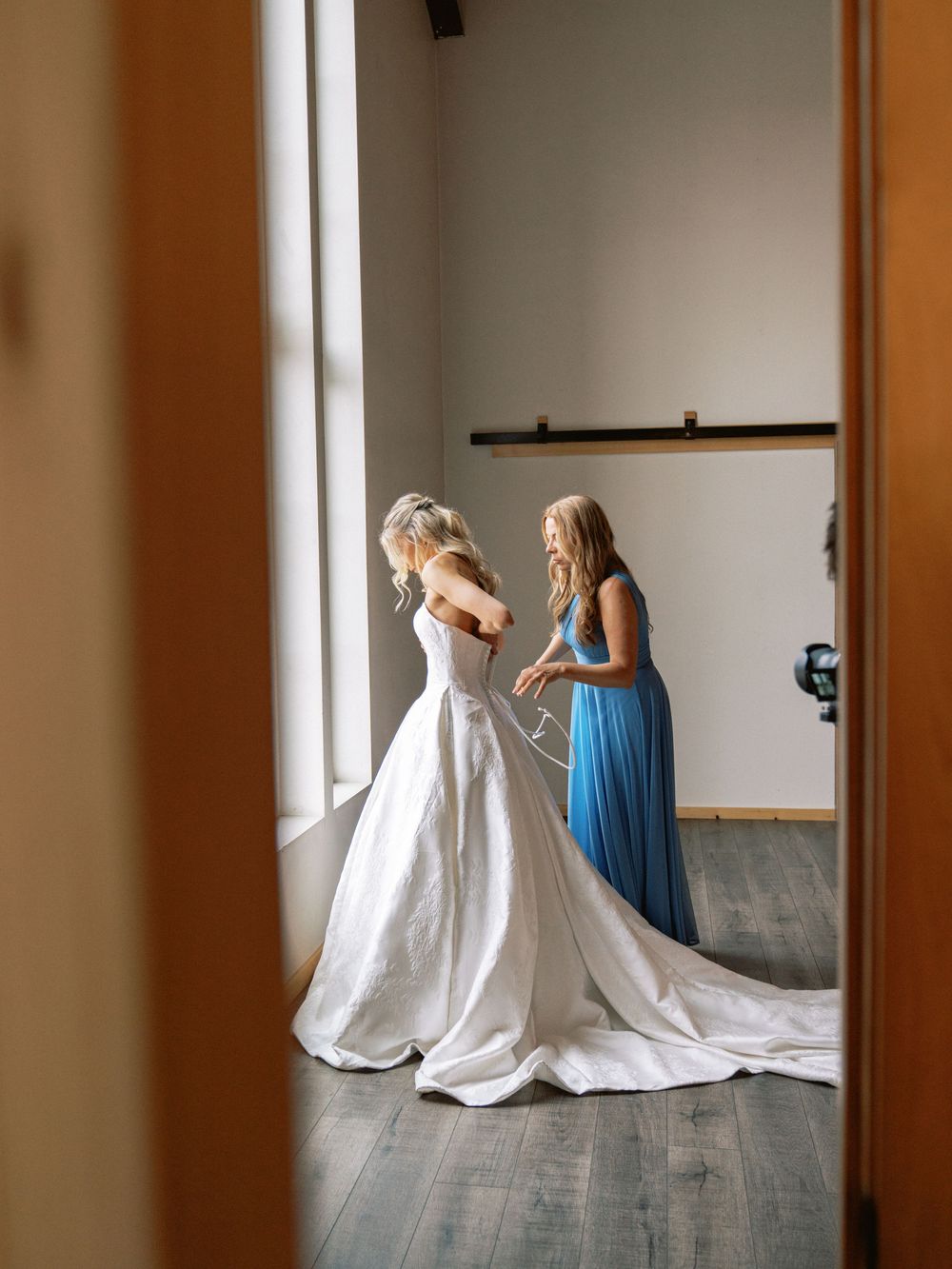 Bridesmaid in blue dress helping bride into white wedding gown in a bright room, viewed through a doorway.