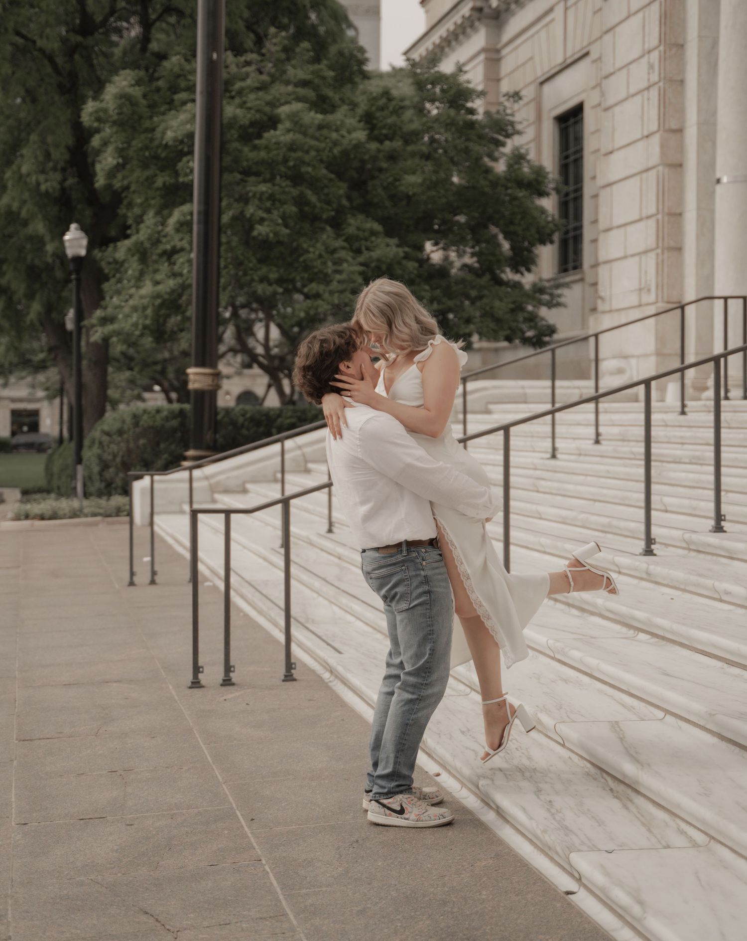 A couple sharing a romantic embrace on marble steps outside a classical building, woman in white dress lifted by man in jeans.