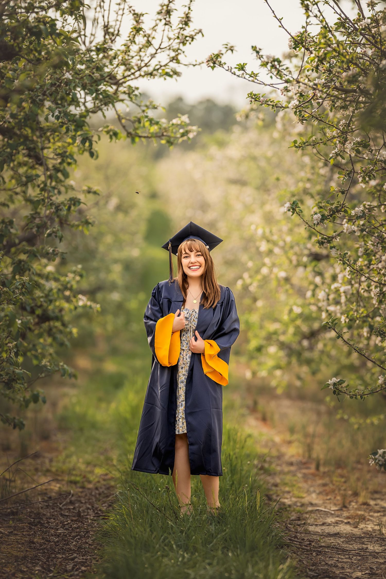 Smiling graduate in navy cap and gown with yellow hood poses on a tree-lined path in bloom.