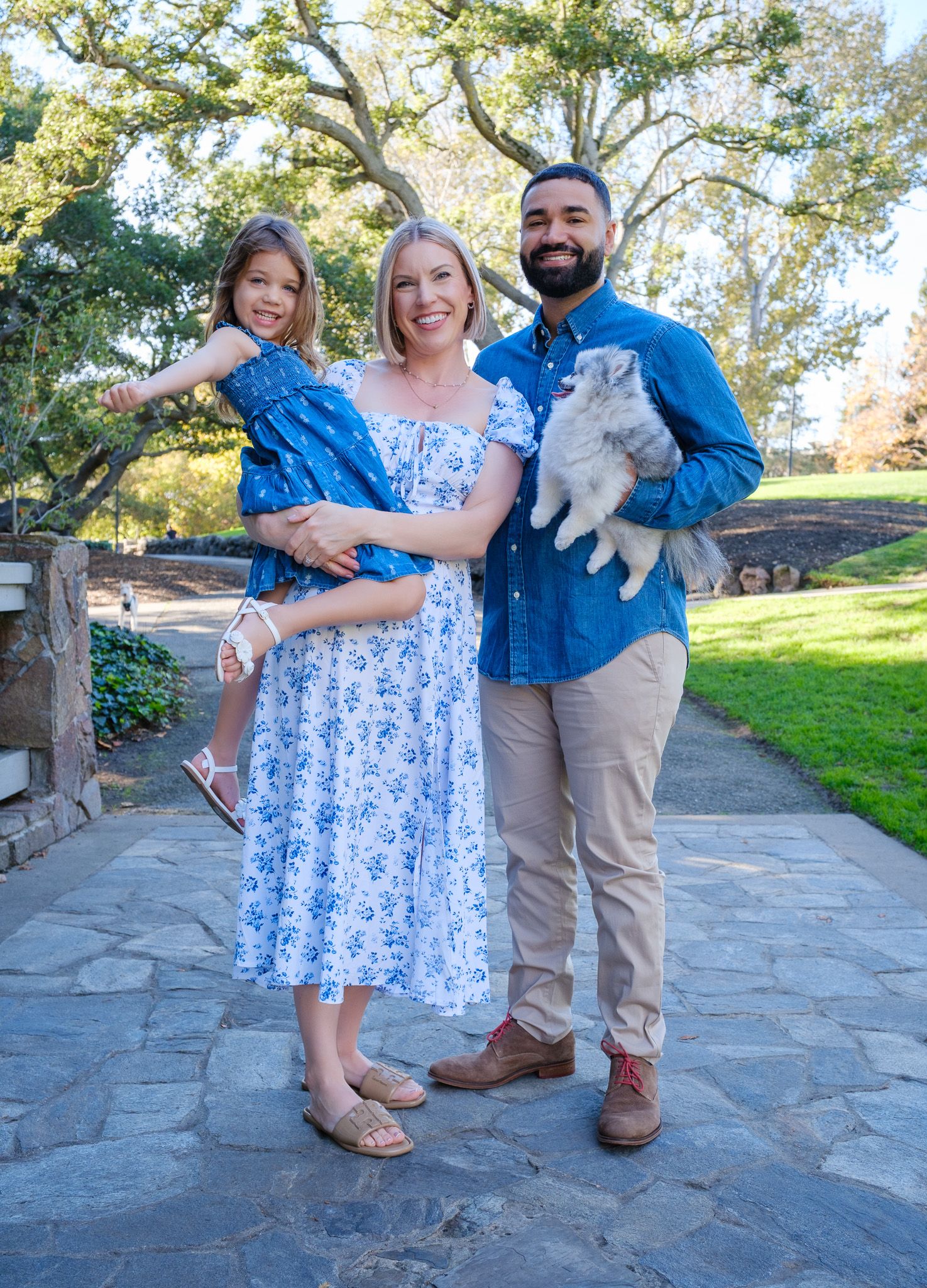 A smiling family poses outdoors, woman in floral dress holds girl in blue, man in denim shirt holds a fluffy cat.