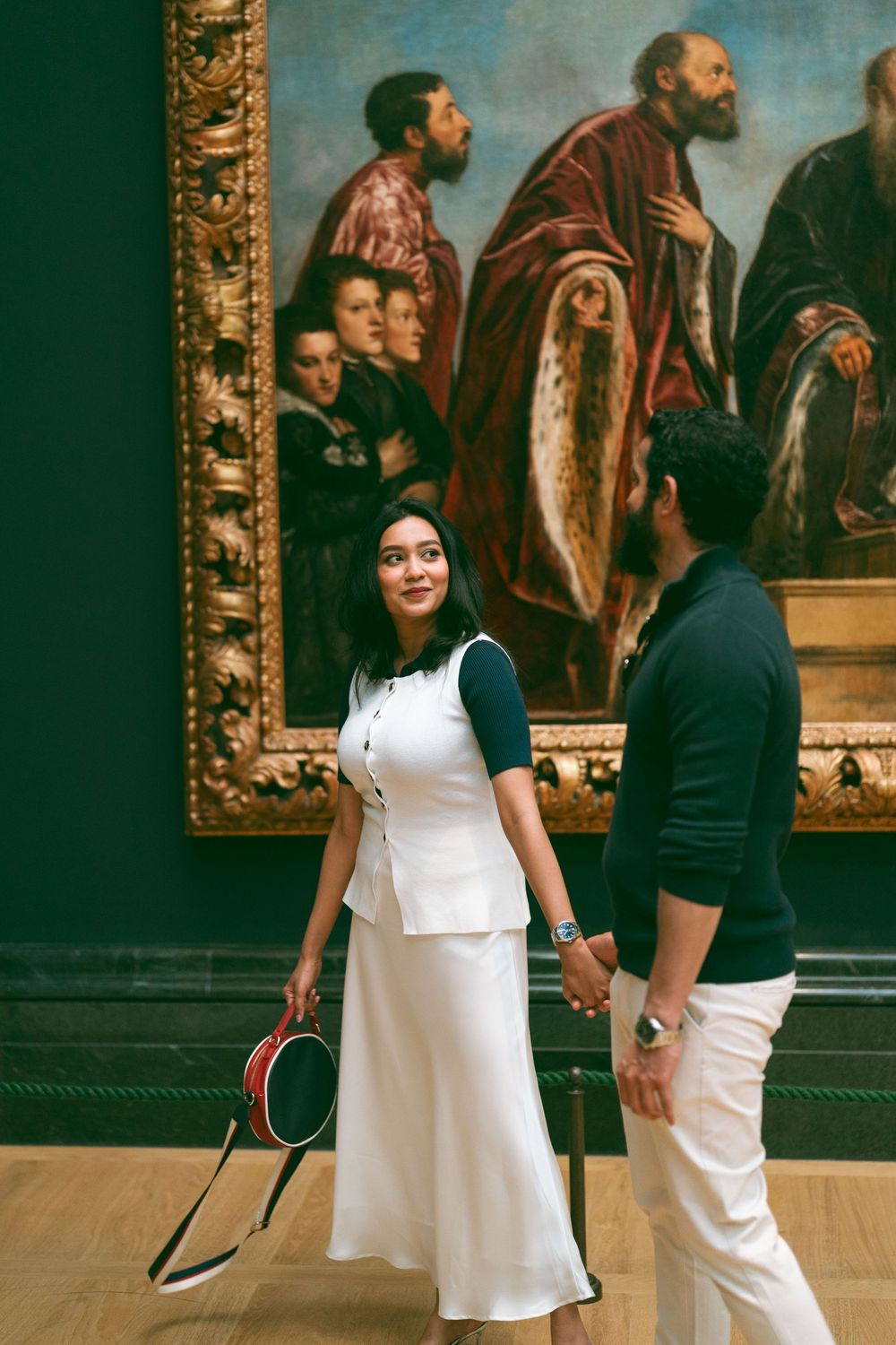 Couple holding hands in art museum, woman in white dress smiling, classic painting displayed on green wall behind them.