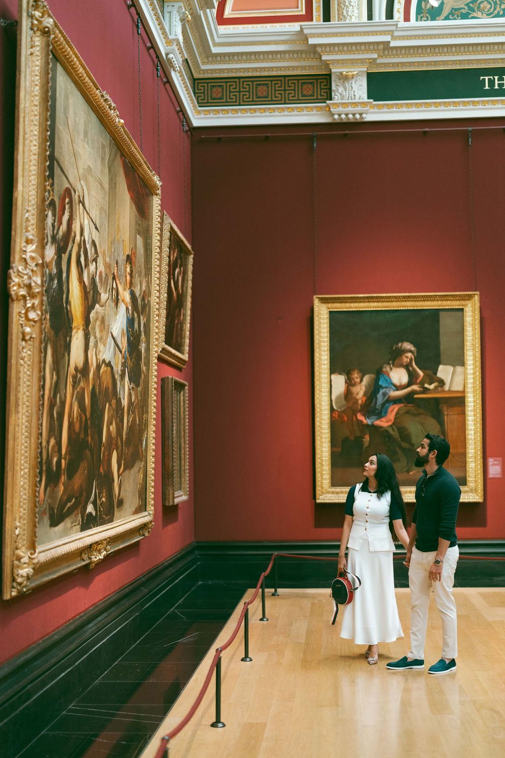 Couple admiring classical paintings in a grand red-walled art museum gallery.