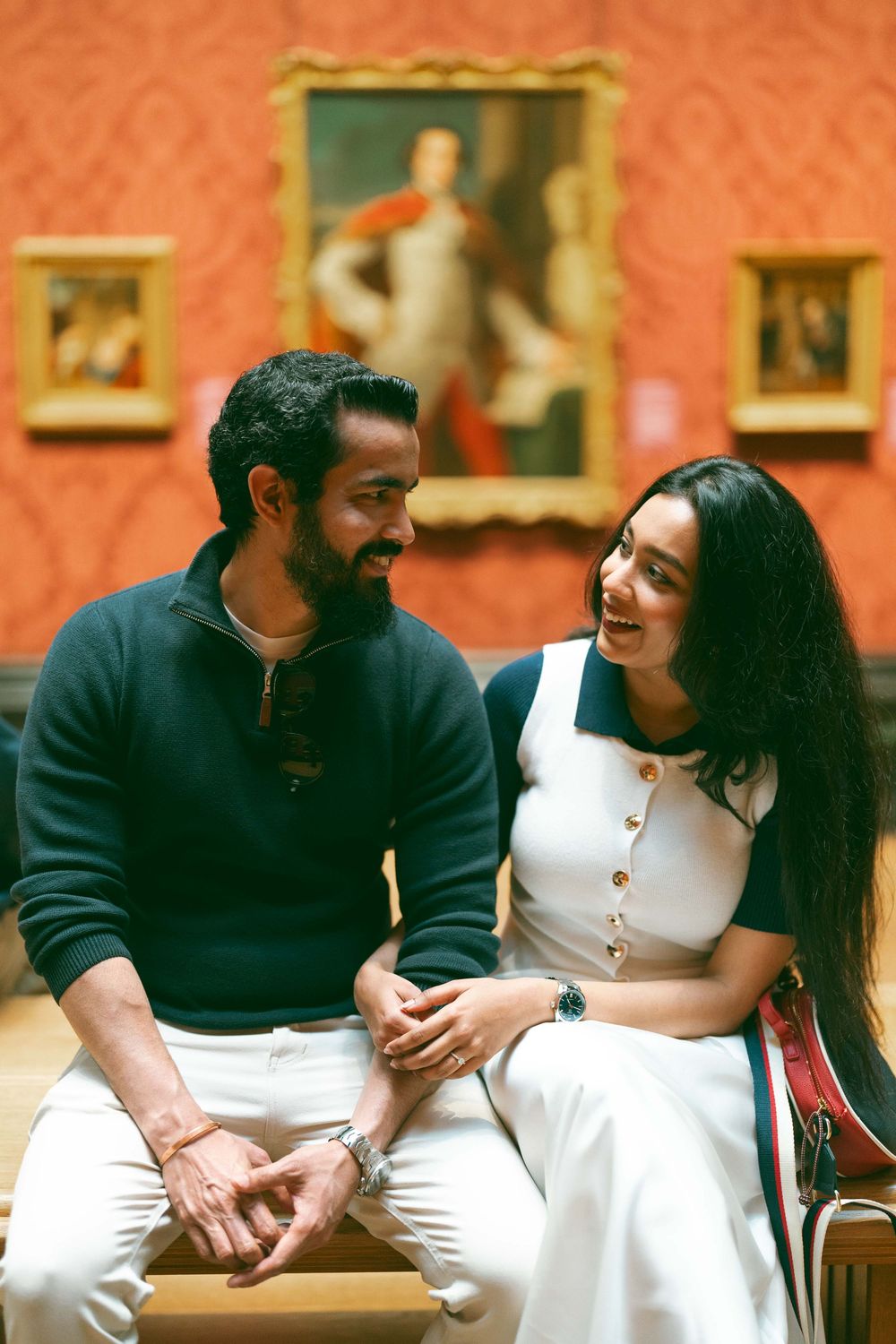 Smiling couple sitting together on a bench in a red-walled art museum gallery.