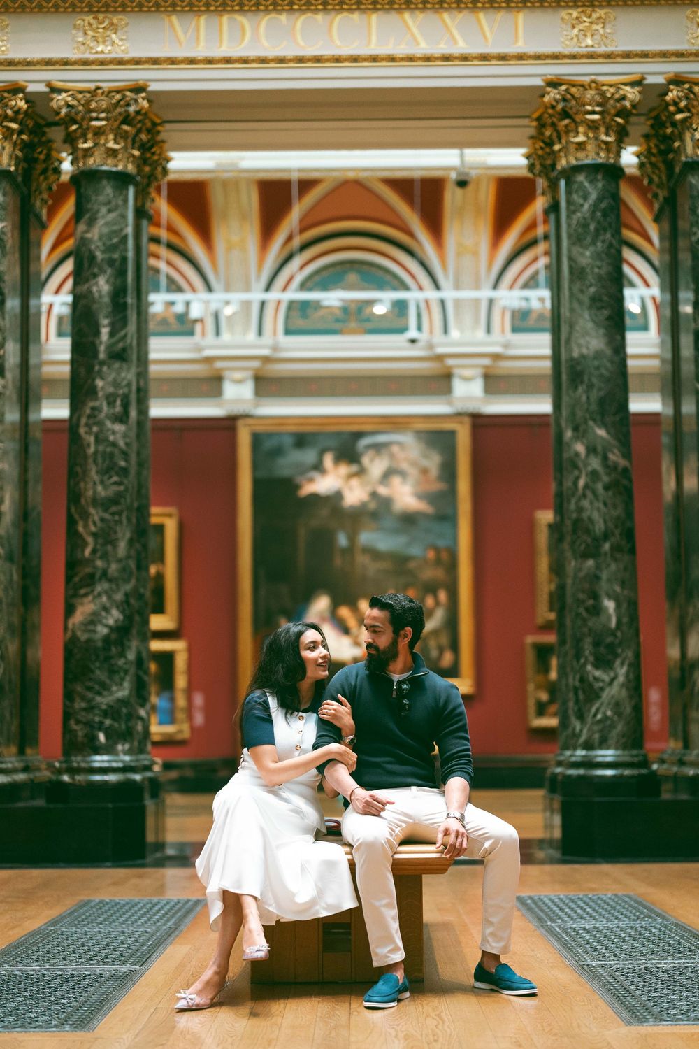 Couple sitting on bench in ornate art museum gallery with classical paintings and marble columns.