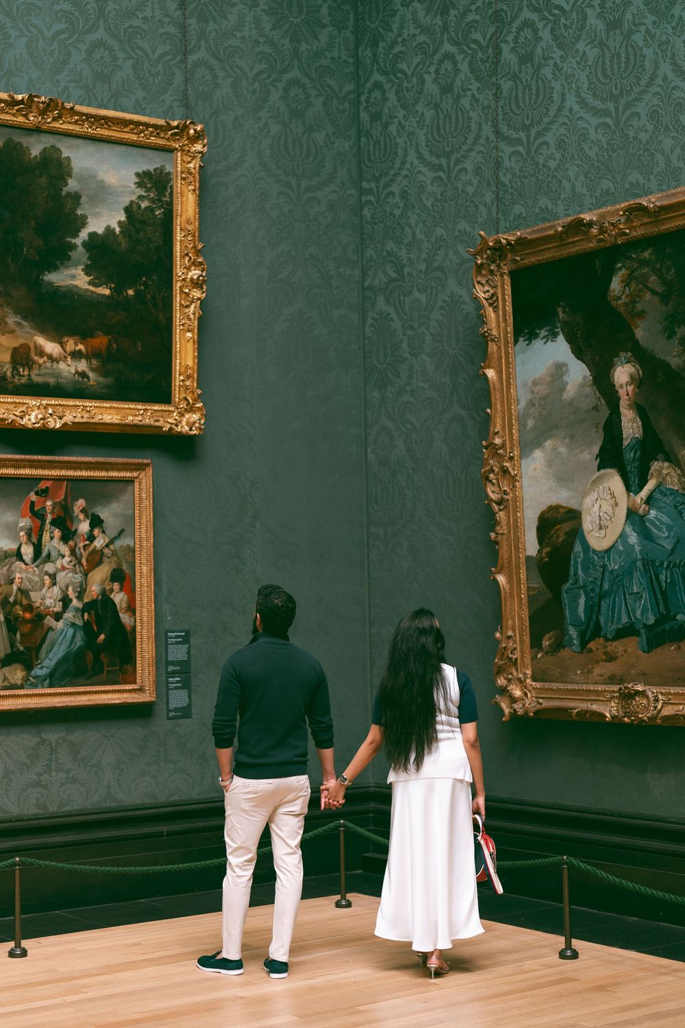 Couple holding hands viewing classic oil paintings in ornate gold frames at an art museum with green damask walls.