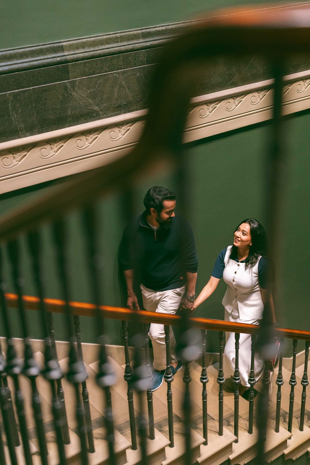 Couple holding hands and smiling on a grand staircase with ornate iron railings indoors.
