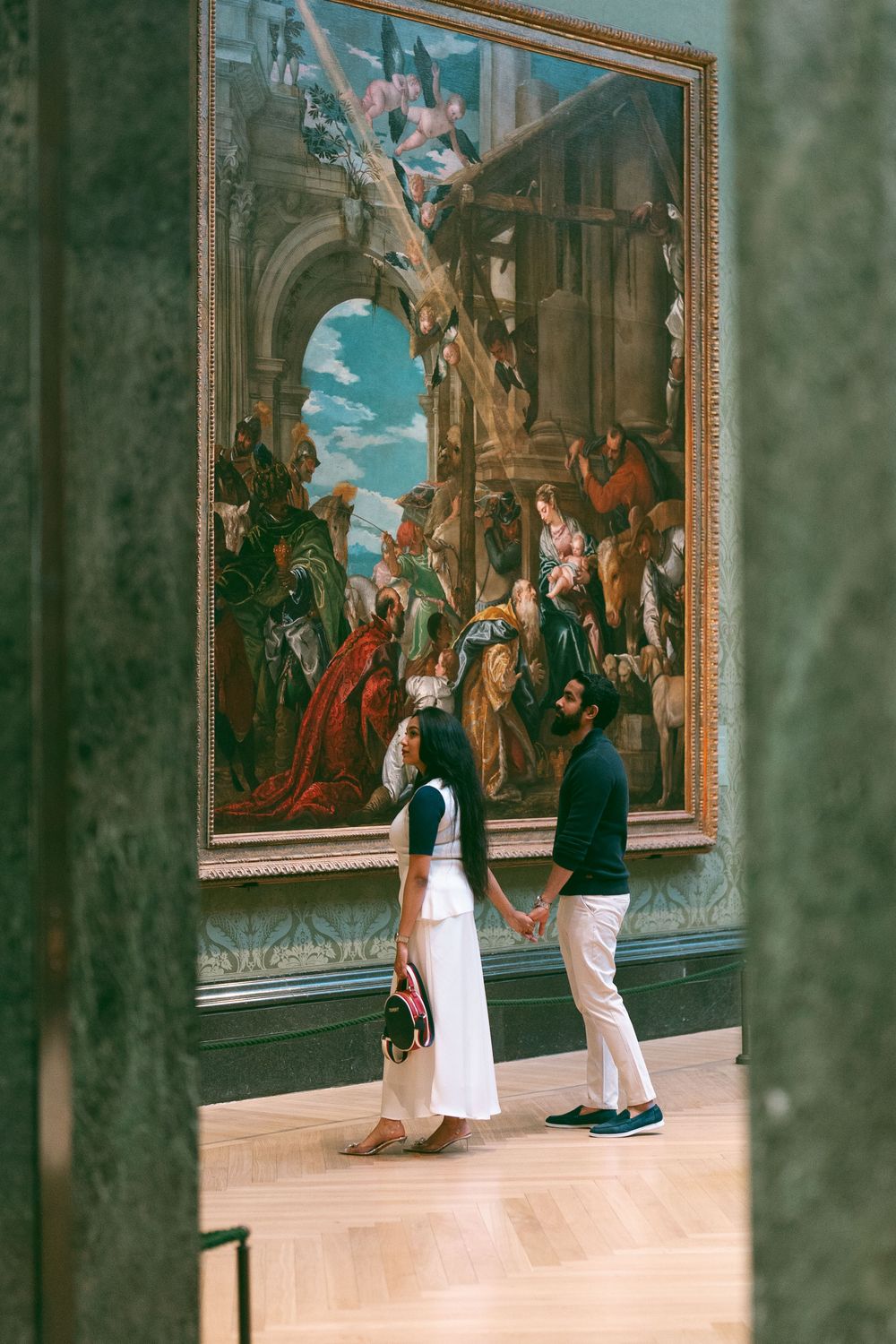 Couple holding hands admiring a large classical painting in an art museum gallery.
