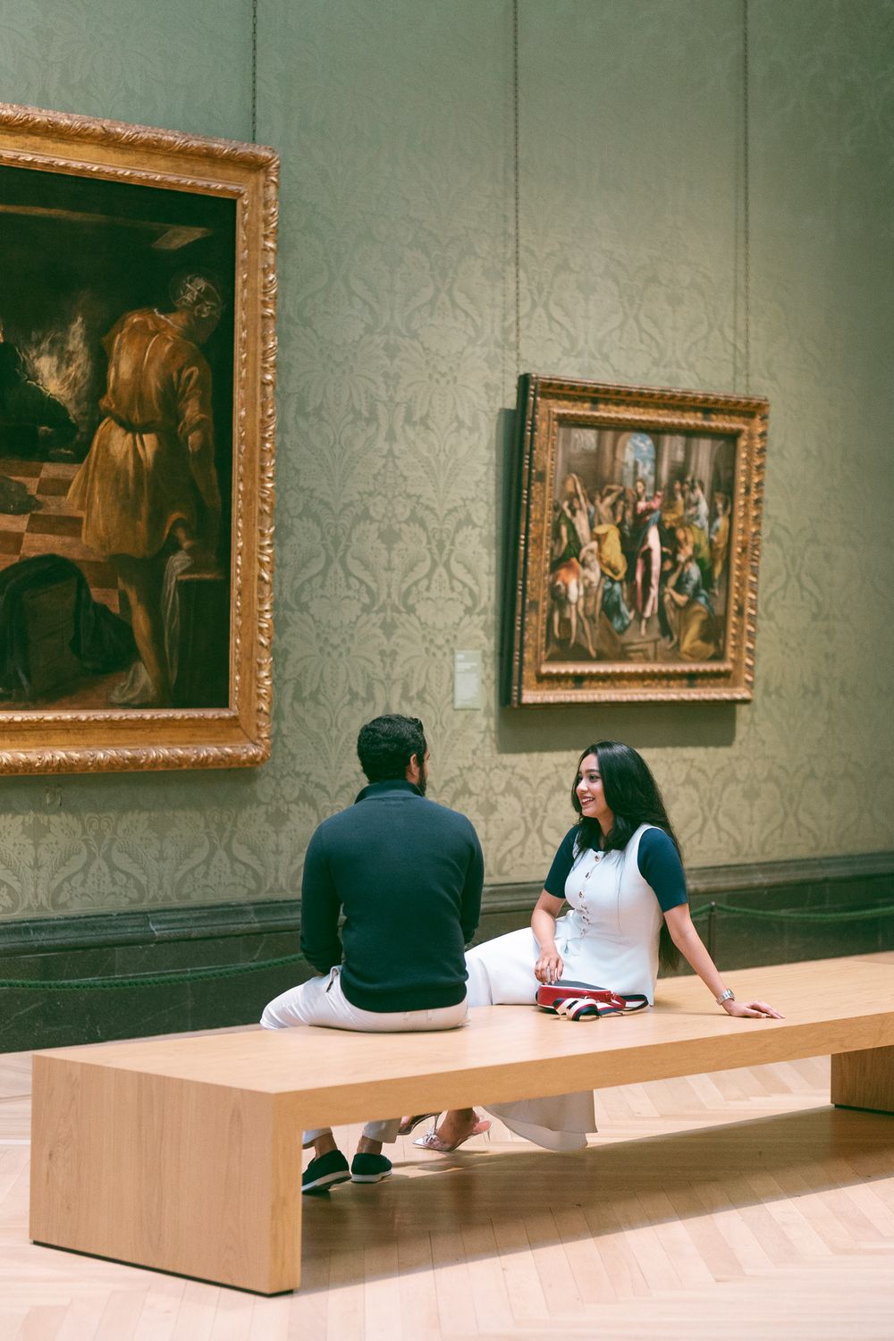 Two people sit on a wooden bench in an art museum, surrounded by large classical paintings on green walls.