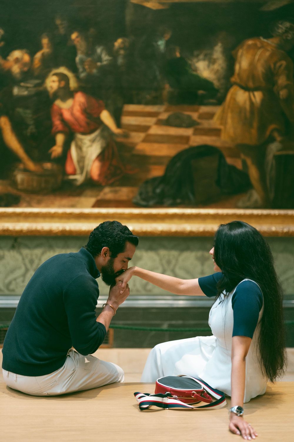 A couple sits on a museum floor, man kissing woman's hand before a large classical painting.