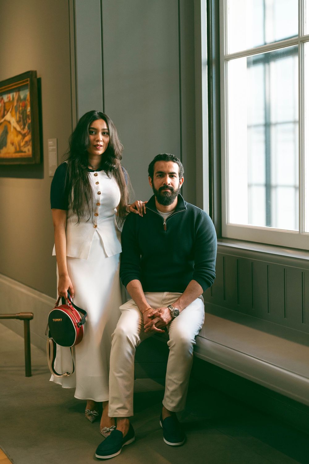 A stylish couple poses in an art gallery, woman in white dress holding red bag, man in navy sweater seated by window.