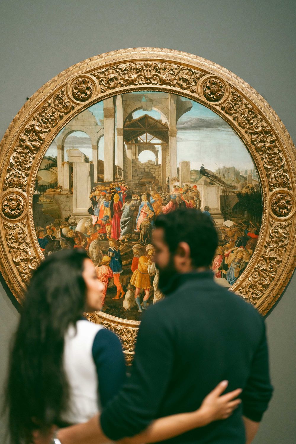 Couple embracing in front of a large circular Renaissance painting in a museum gallery.