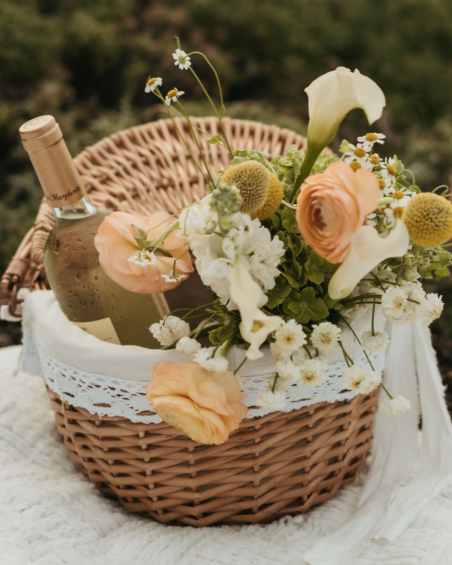 Wicker picnic basket with peach roses, white calla lilies, chamomile flowers, and a wine bottle on lace cloth.