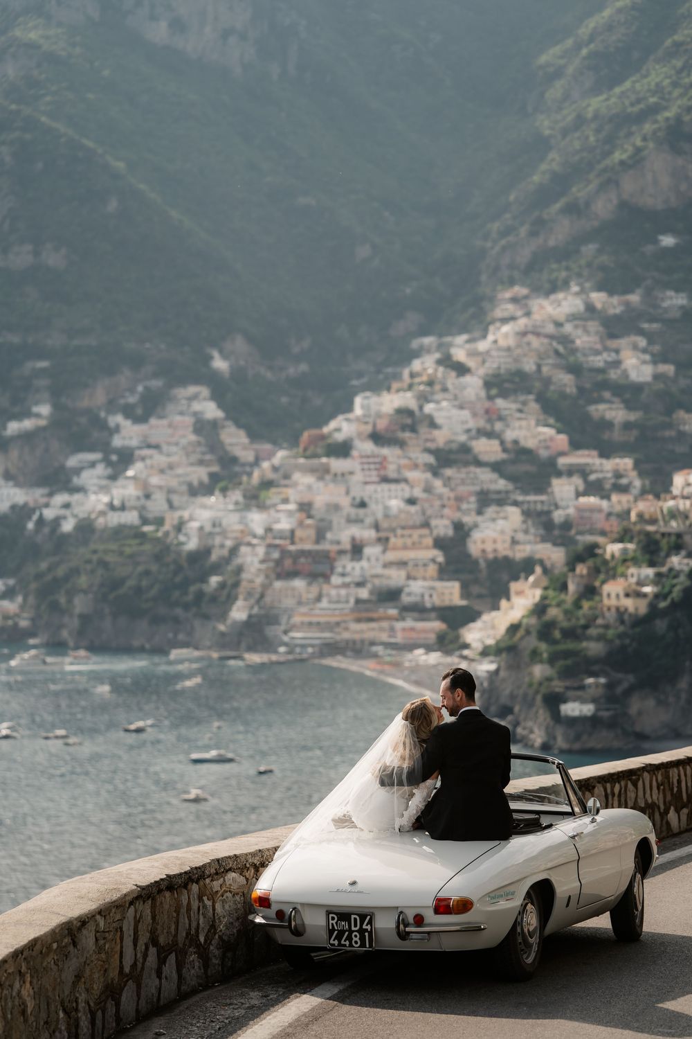 Bride and groom kissing in a white vintage convertible on the Amalfi Coast with Positano village backdrop.