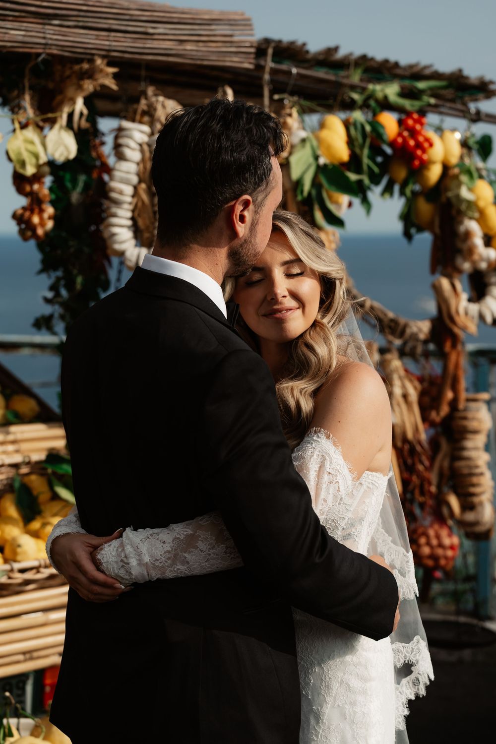 Bride and groom embracing at outdoor wedding ceremony with rustic fruit and floral arch near the ocean.