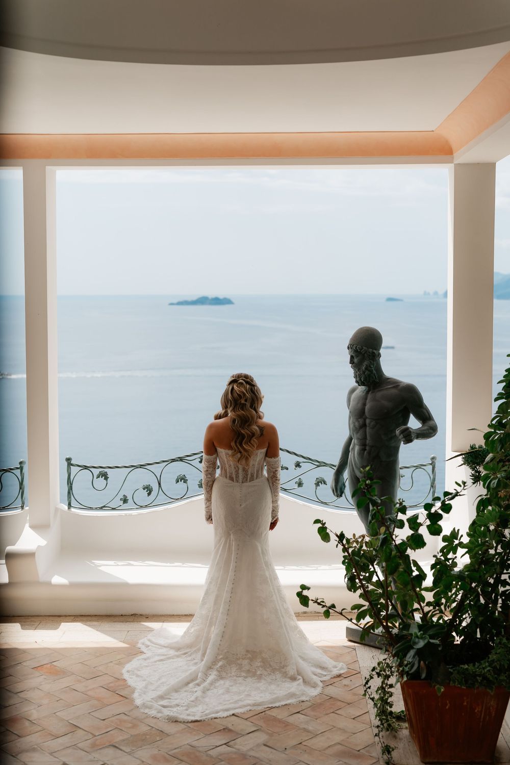 Bride in lace gown stands before ocean view balcony beside bronze male statue at elegant coastal venue.