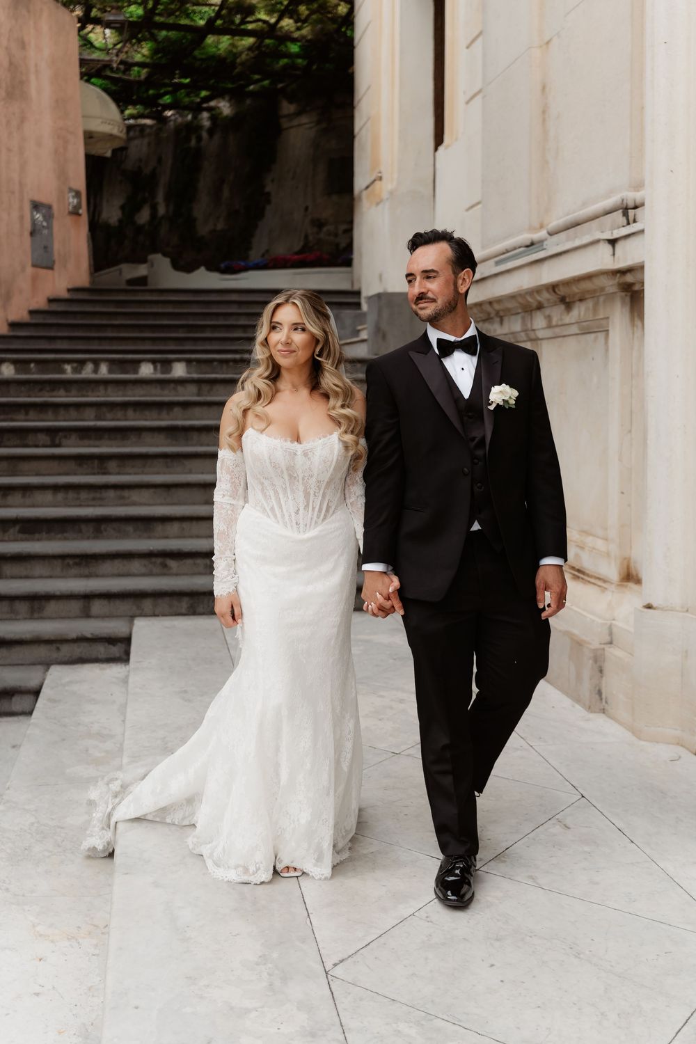 A bride in a white lace gown and groom in a black tuxedo hold hands walking near stone steps.