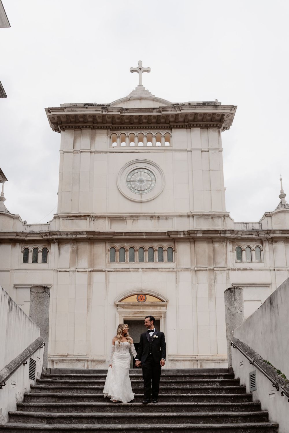 Bride and groom standing on church steps in front of a white historic chapel with a cross on their wedding day.