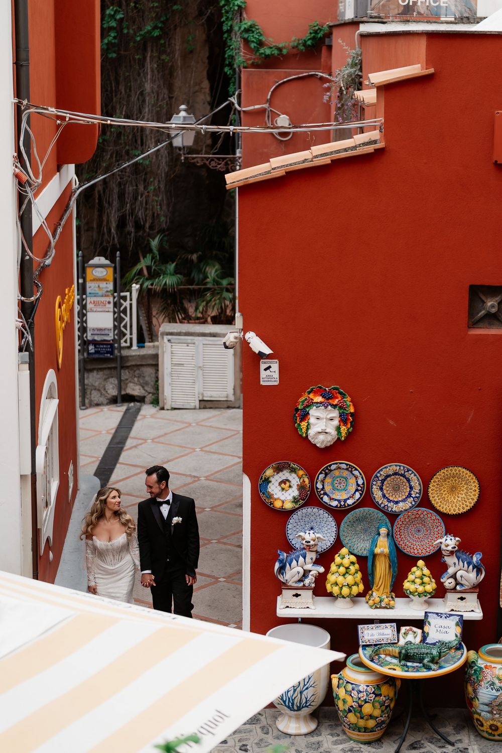 Bride and groom walking in a colorful Italian alley with vibrant ceramic plates displayed on a red wall in Positano.