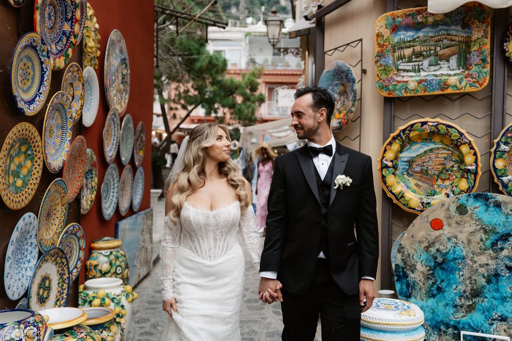 Bride and groom hold hands among colorful Italian ceramic plates displayed in a Positano shop alley.