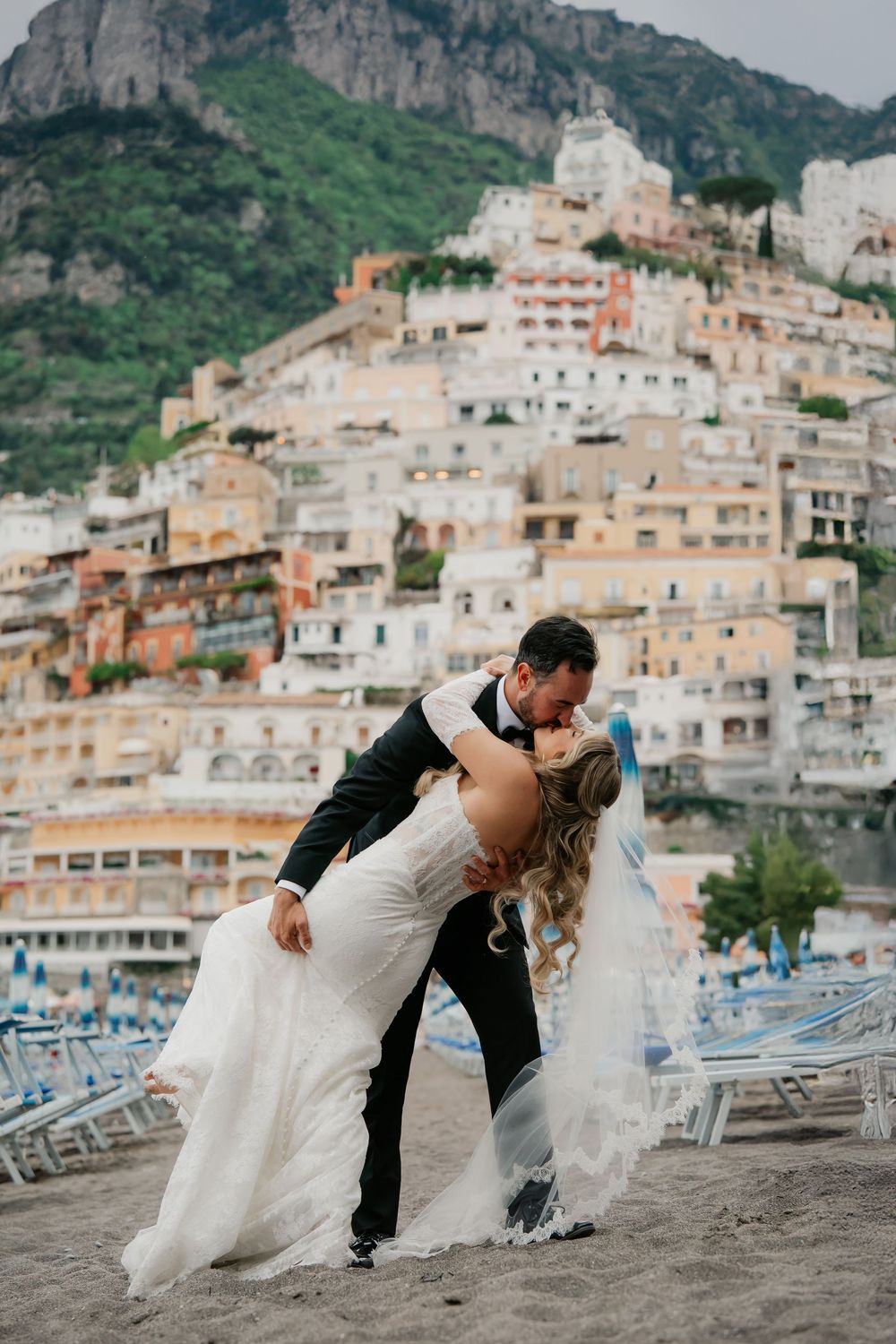 Bride and groom share a romantic dip kiss on Positano beach with colorful cliffside buildings in the background.