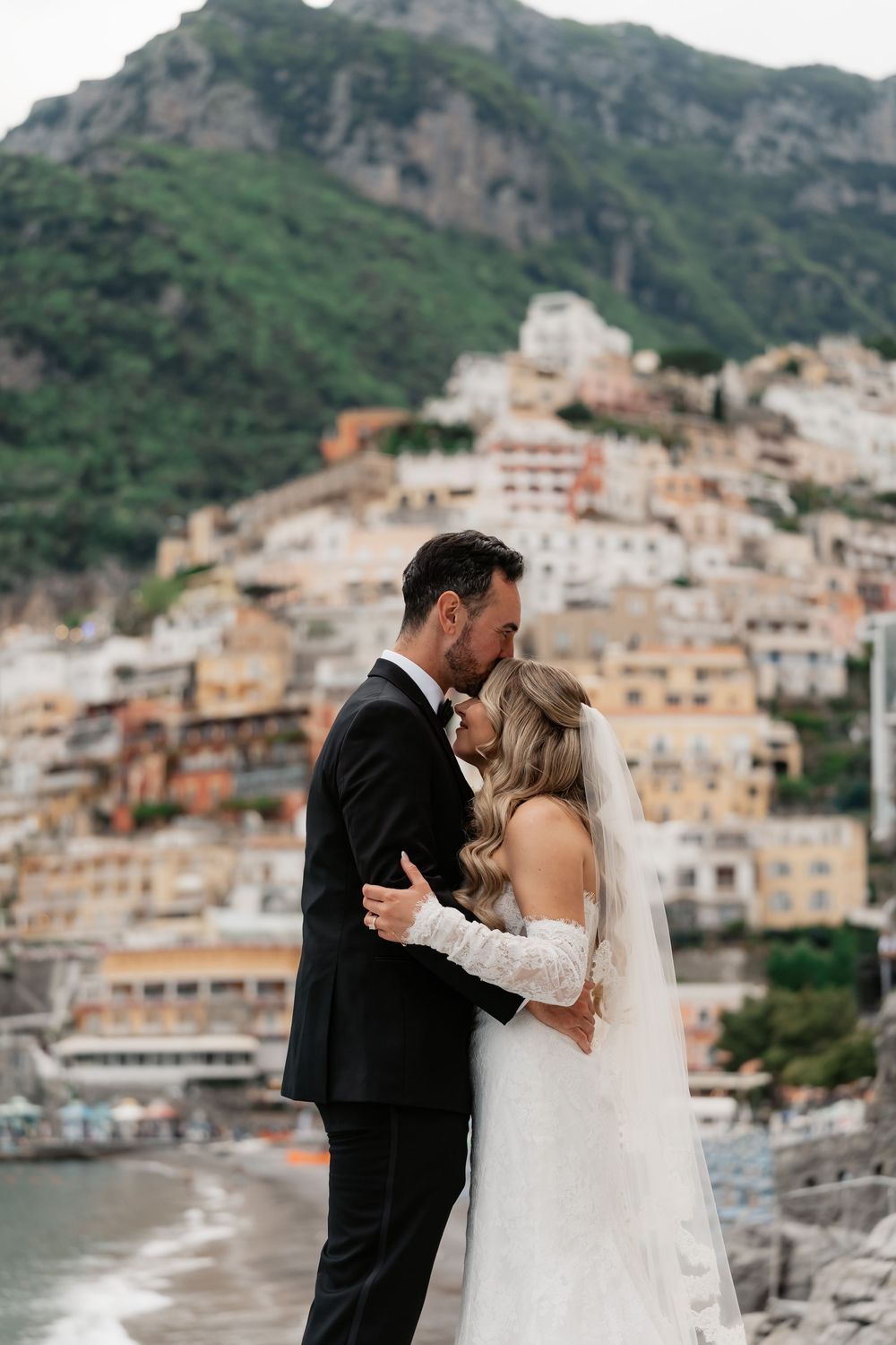 Bride and groom share a romantic kiss with Positano's colorful cliffside village and mountains in the background.