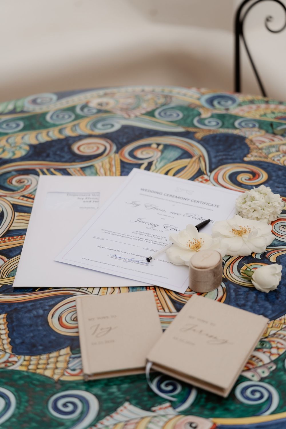 Wedding ceremony certificate with pen, white flowers, and vow books on decorative mosaic table.