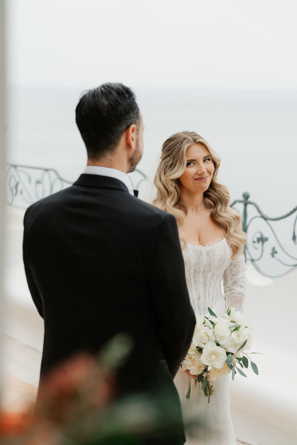 Bride in lace gown holding white bouquet smiles at groom in black tuxedo during outdoor wedding ceremony by the sea.