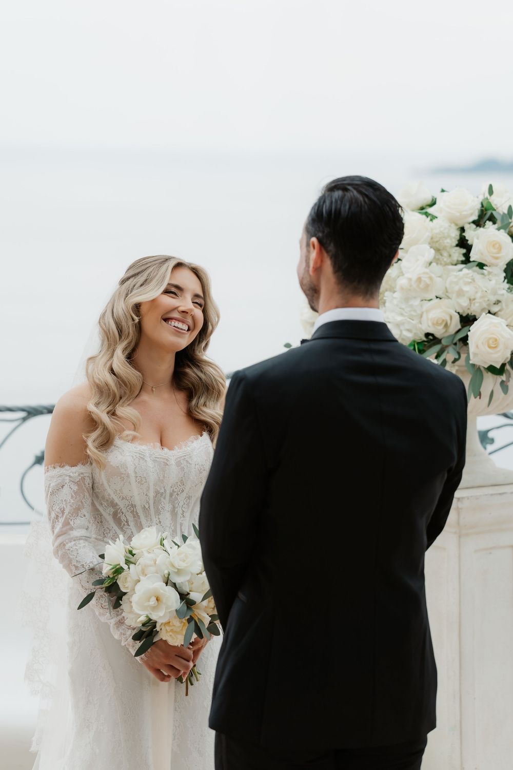 Smiling bride in lace off-shoulder gown holds white bouquet facing groom in black tuxedo at outdoor wedding ceremony.