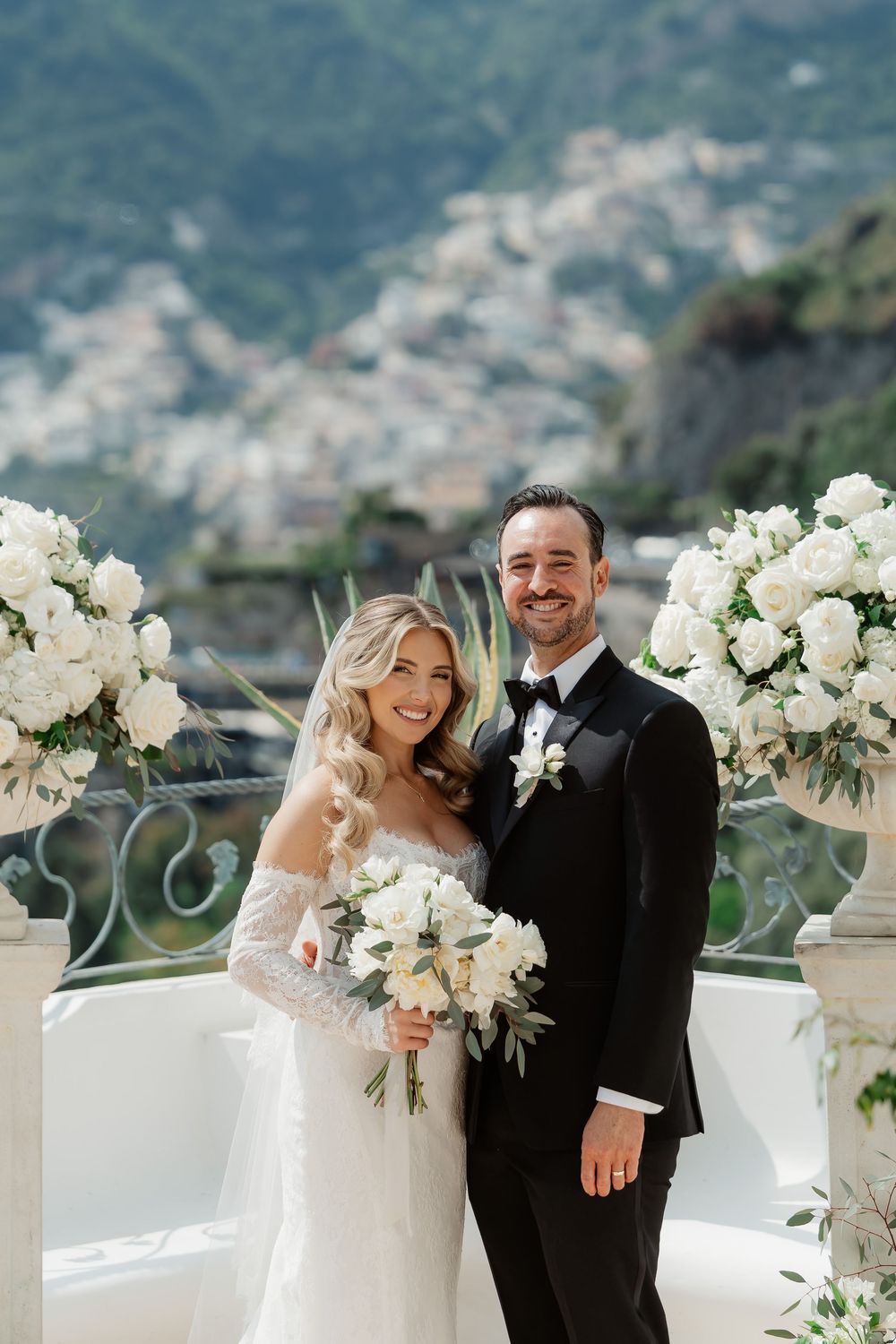 Bride and groom smiling on elegant terrace with white floral arrangements and Positano hillside backdrop.