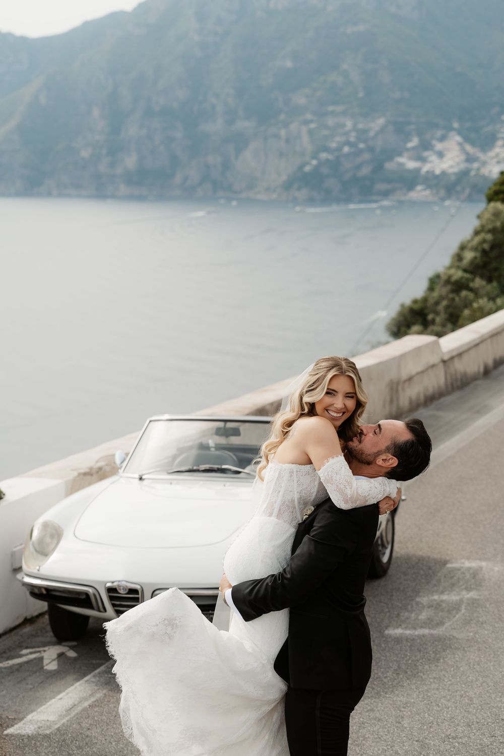 Groom lifts smiling bride in white gown beside vintage convertible on Amalfi Coast with scenic sea views.