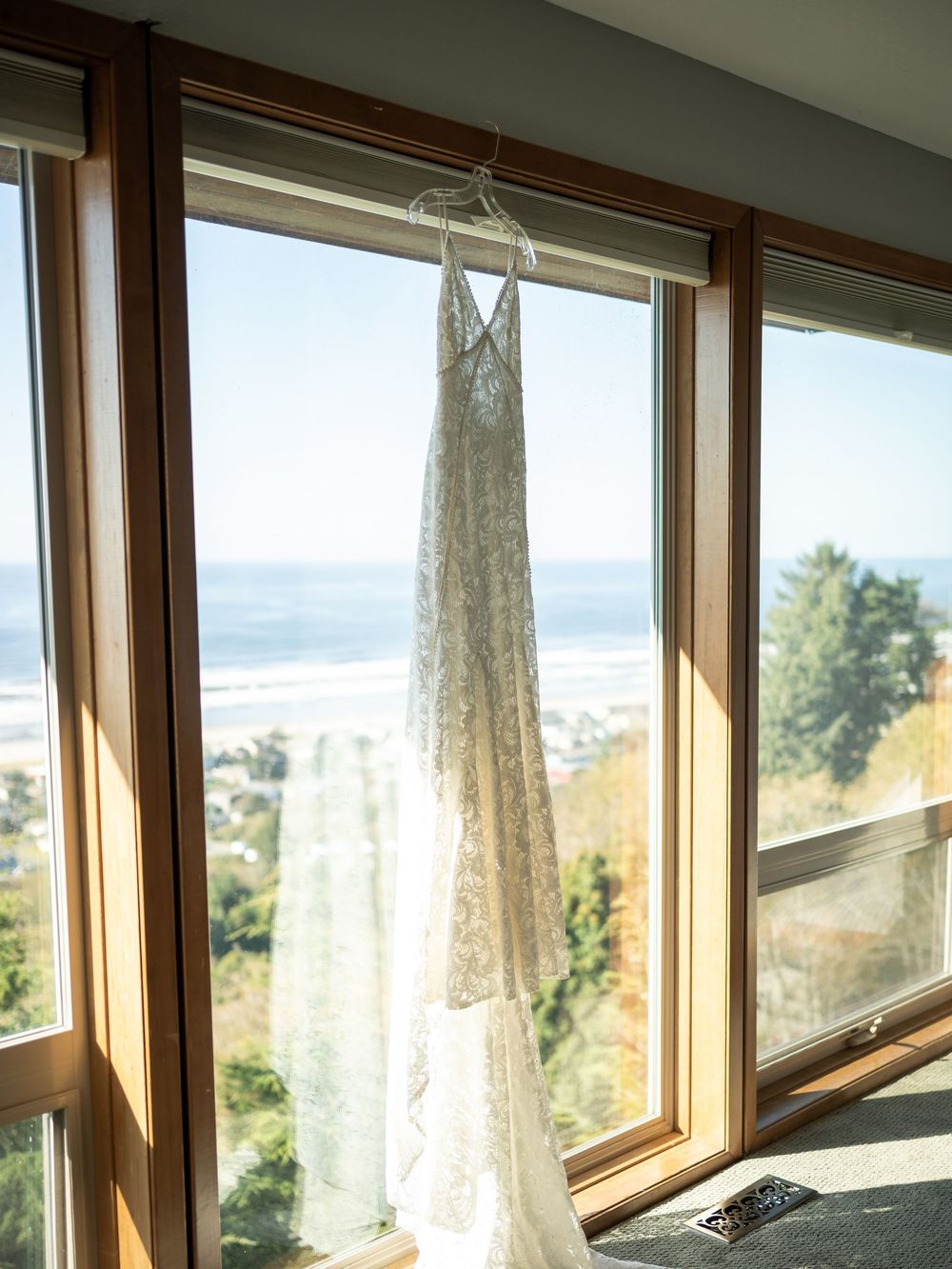 Lace wedding dress hanging on window with ocean view backdrop on sunny day.
