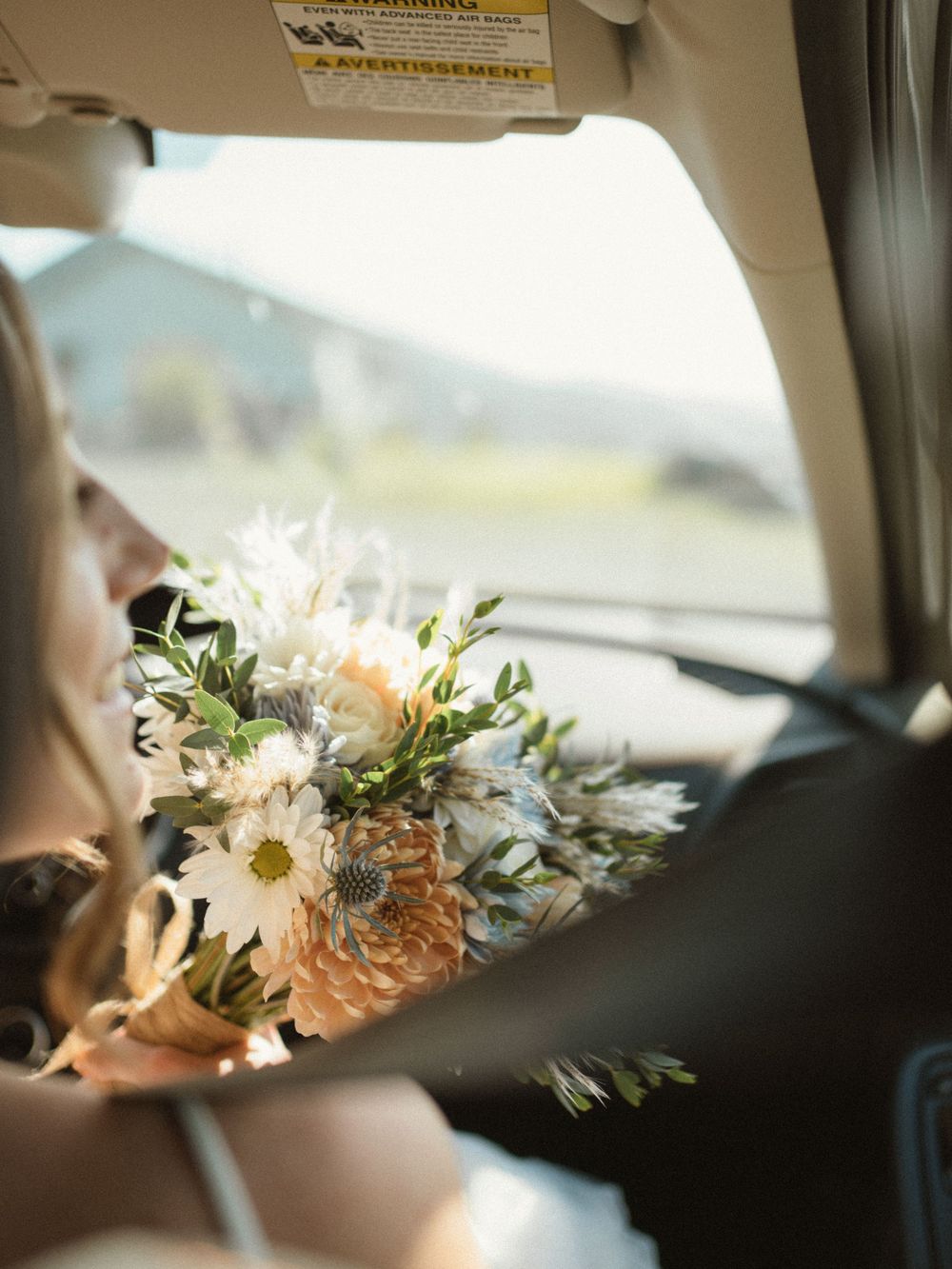 Bride holding a white and peach wedding bouquet with daisies while riding in a car on her wedding day.