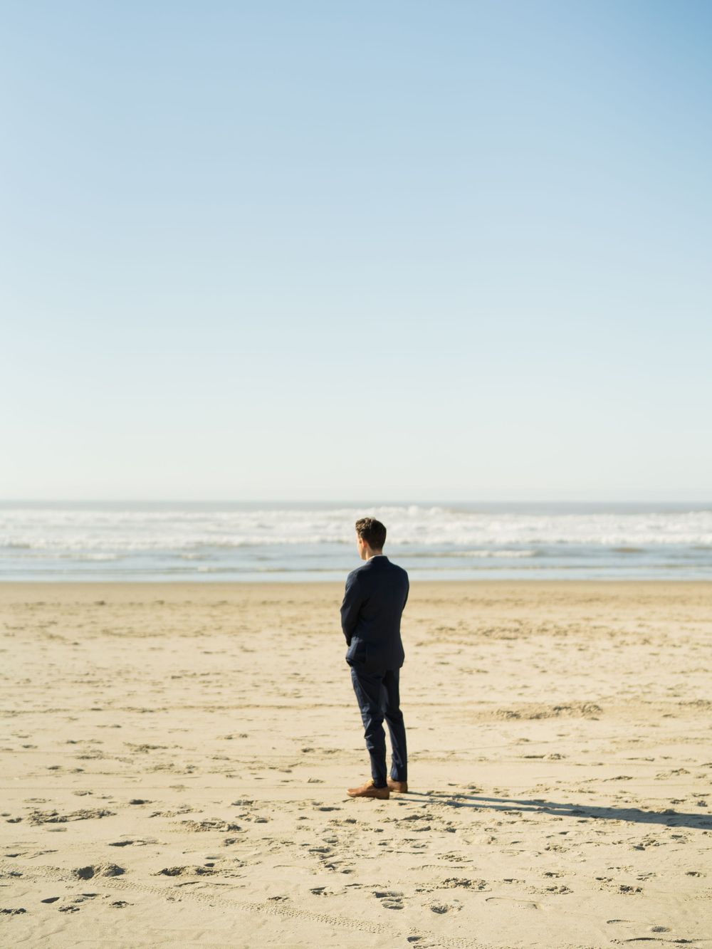 Groom in navy suit stands alone on sandy beach gazing at ocean horizon on clear sunny day.