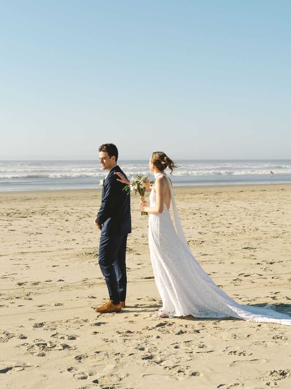 Bride and groom first look on sandy beach with ocean waves and clear blue sky.