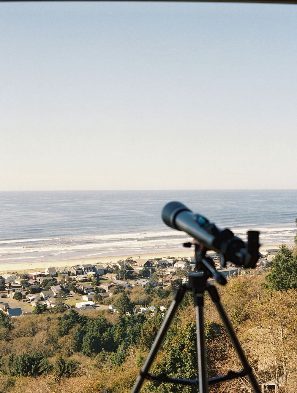 Telescope on tripod overlooking a scenic coastal town and sandy beach on a clear sunny day.