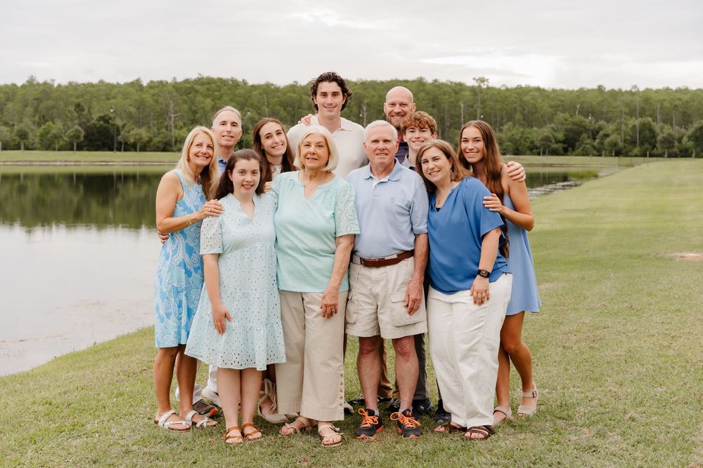 Multi-generational family of ten posing together outdoors near a serene pond surrounded by lush green trees.