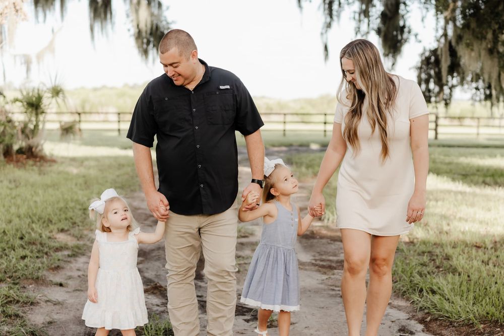A happy family of four walks hand-in-hand outdoors, with two young girls in white and blue dresses on a sunny day.