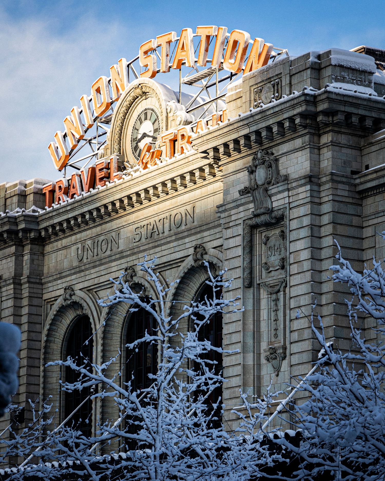 Snow-dusted Union Station in Denver with glowing neon sign and frost-covered trees under a blue winter sky.