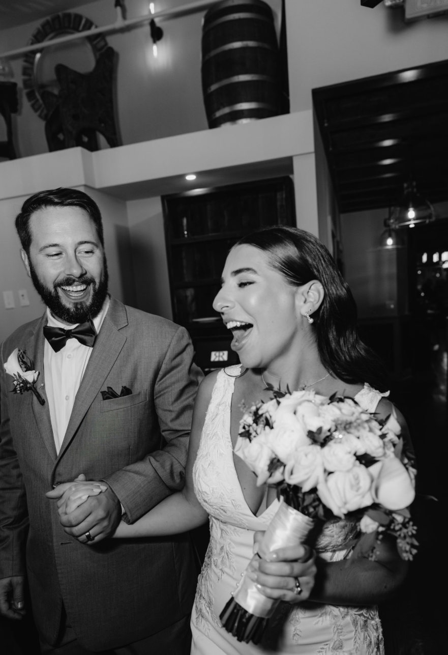 Black and white photo of a joyful bride holding bouquet and groom in bow tie laughing together at wedding reception.