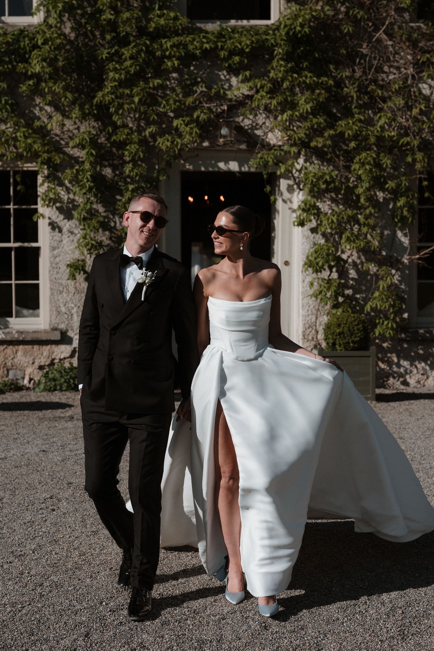 Stylish bride and groom walking outside ivy-covered venue, wearing sunglasses, black tuxedo and strapless white wedding gown.